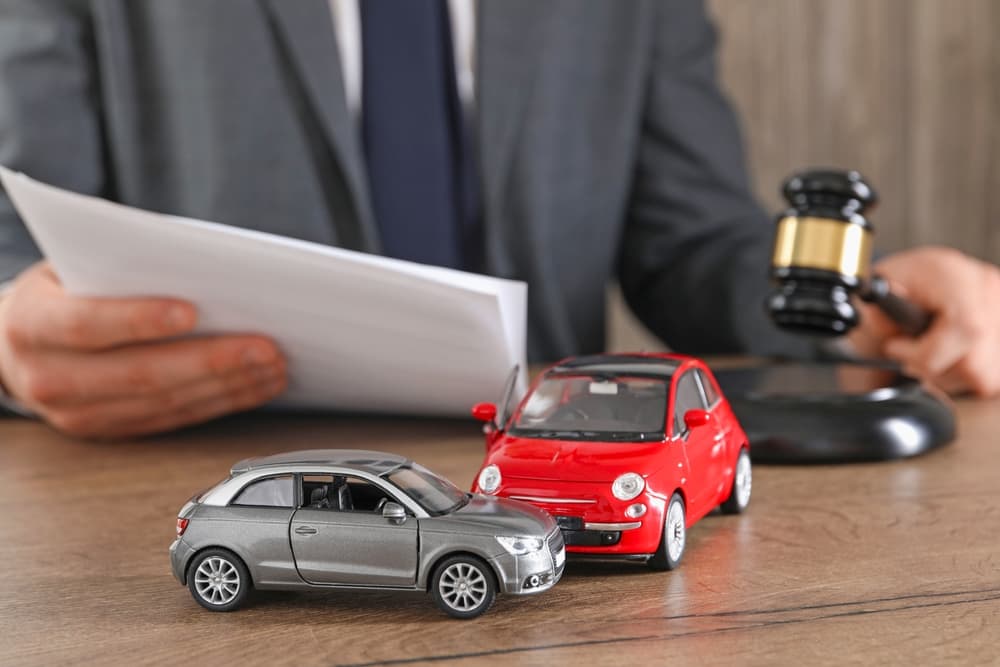 A man with documents and a gavel at a wooden table indoors, with focus on toy cars representing the incident.