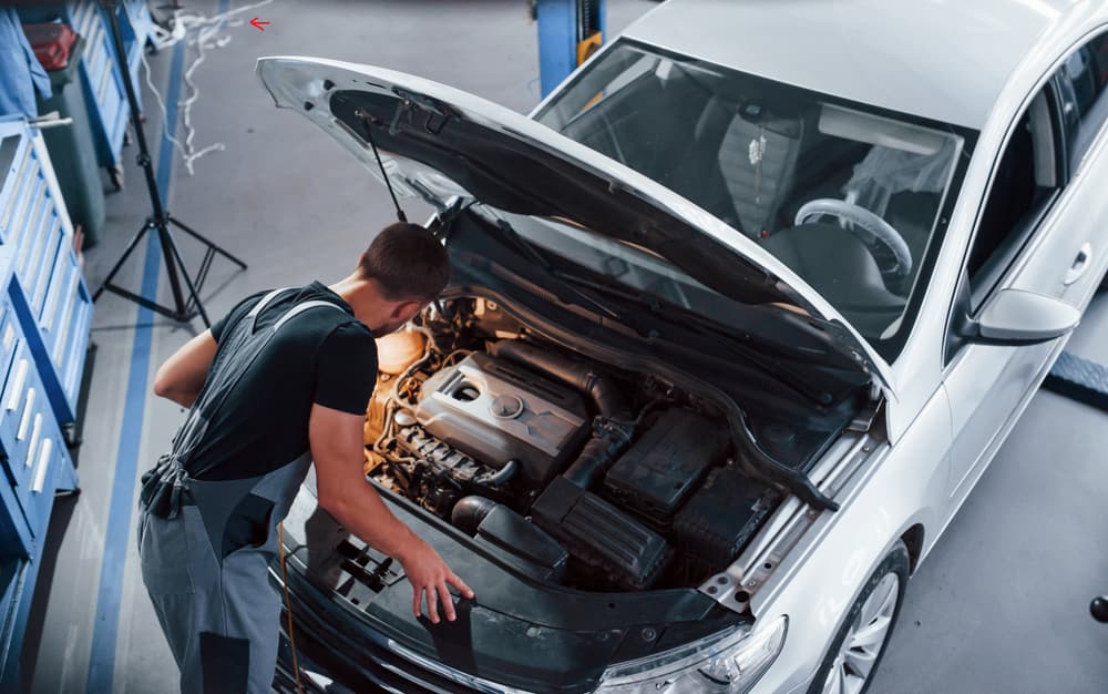 Under the hood: A man in a grey uniform repairs a white automobile indoors.