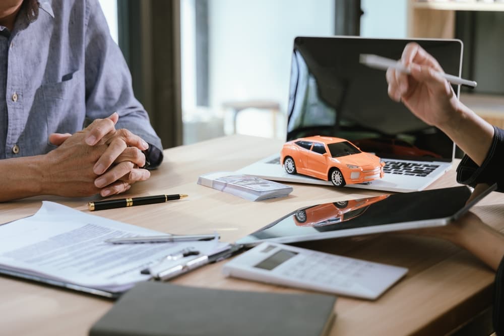 A car salesman and a customer negotiate a deal in an office. Both smile as the customer signs the car insurance contract.