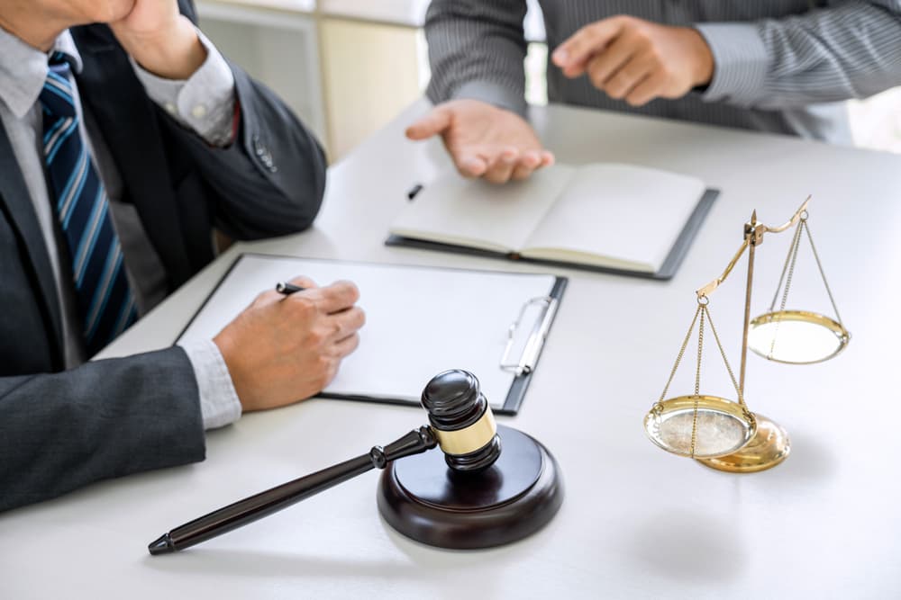 At a law firm office, a businessman and lawyer consult and discuss contract papers, accompanied by a judge’s gavel and scales of justice symbolizing legal authority.