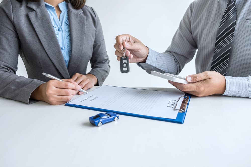 A woman signs a car insurance document or lease agreement, writing her signature on the contract, while a man hands her the car keys.