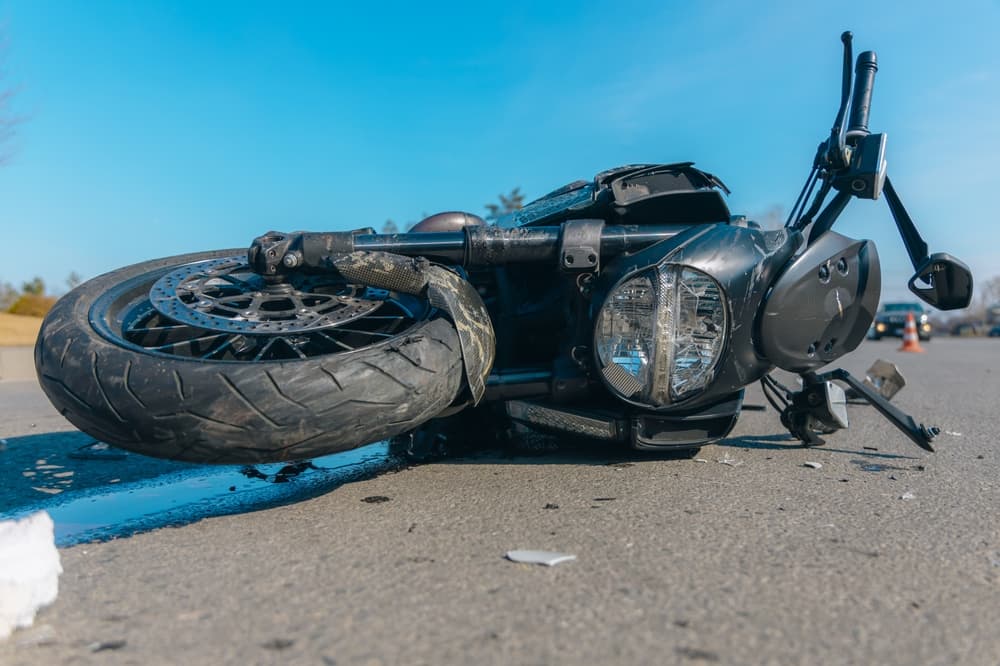 A black motorcycle lies severely damaged on the asphalt after an accident. Close-up shows a broken headlight and bent handlebars, indicating a loss of control or a collision with another vehicle.
