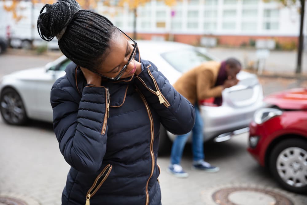 Woman suffering from neck pain while a man examines a damaged car on the road.