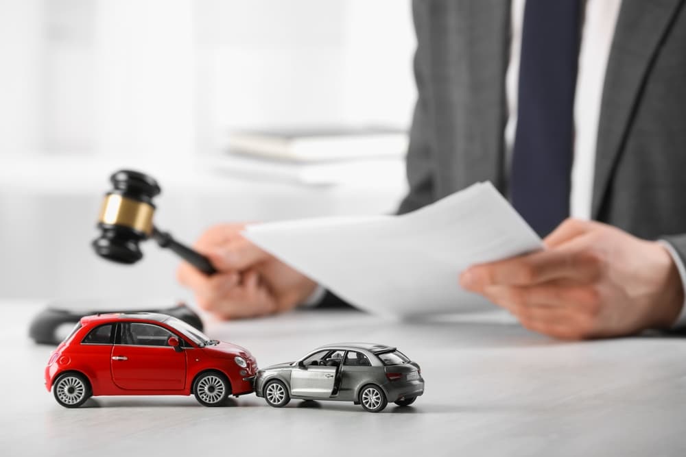The image illustrates auto accident law, featuring a lawyer with legal documents and a gavel at a table indoors, with the focus on toy cars symbolizing the collision.