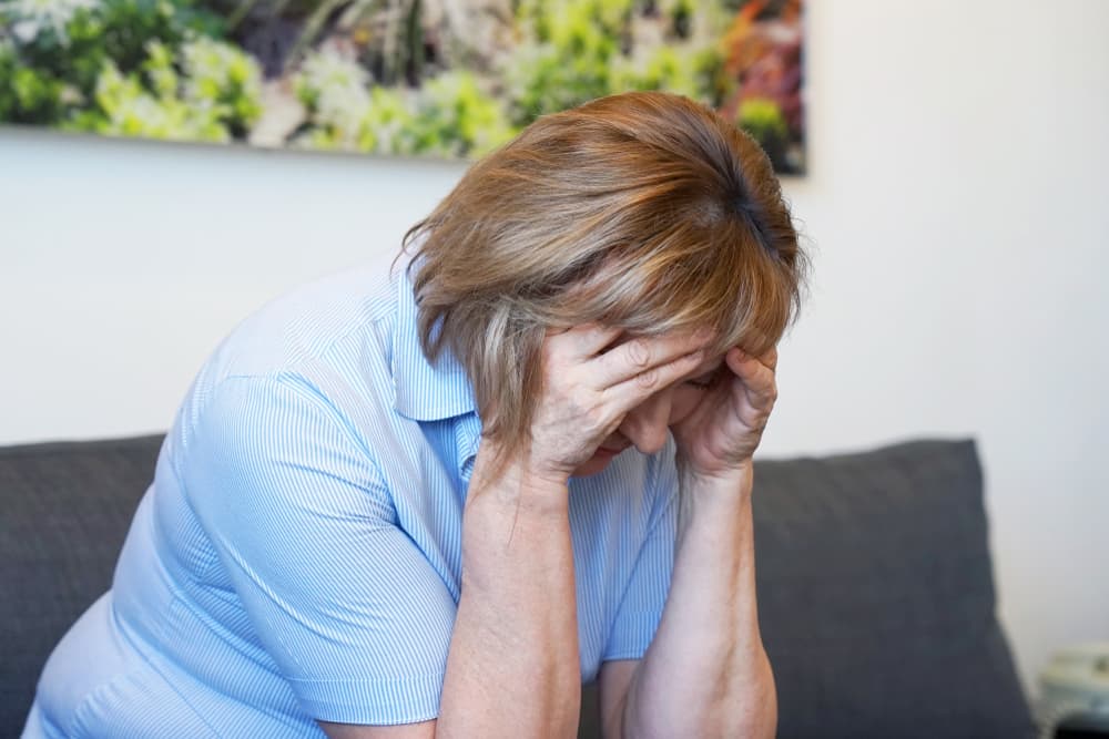 An elderly woman sits at home holding her head in pain, suffering from a headache or migraine, representing issues related to health, stress, and depression.