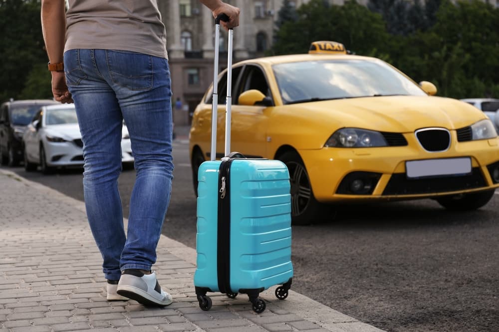 A close-up image shows a man with a suitcase standing next to a taxi car outdoors, suggesting travel or a recent arrival.
