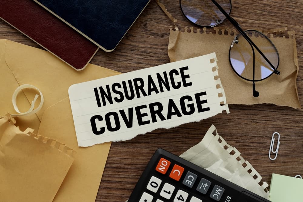 The image shows the words "Insurance Coverage" on torn paper, placed among various stationery items, symbolizing the complexity and paperwork involved in insurance matters.