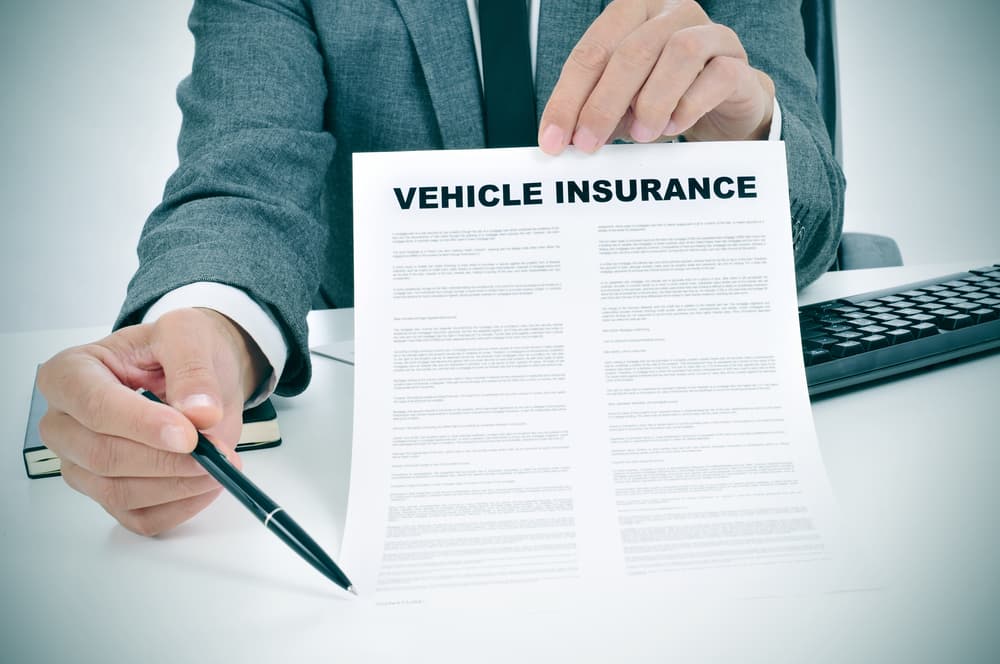 A young man in a gray suit sits in his office, holding a vehicle insurance policy and pointing with a pen to the spot where the policyholder needs to sign.