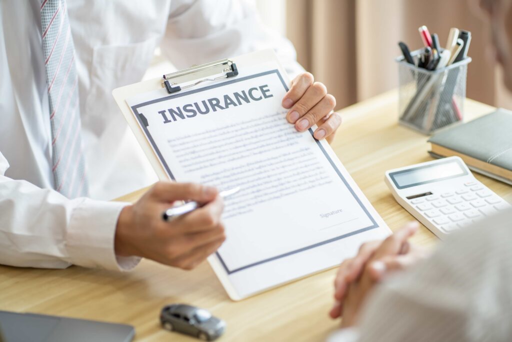 Insurance agent explaining policy document to client with contract, calculator, and paperwork on office desk.