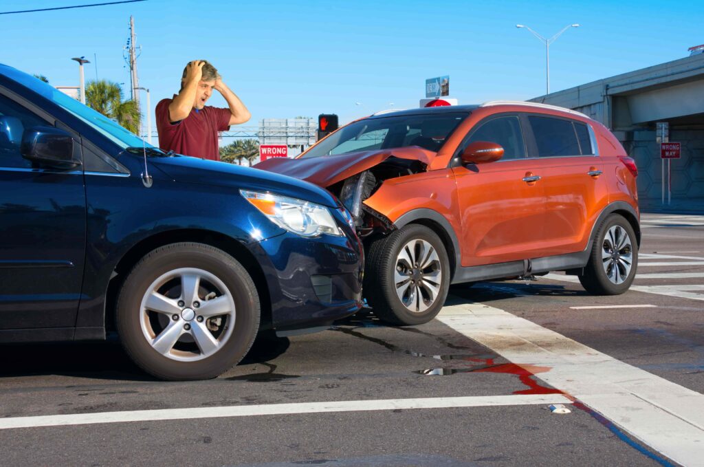 Two-car accident at an intersection with a distressed driver holding his head after a collision between a blue SUV and an orange crossover.