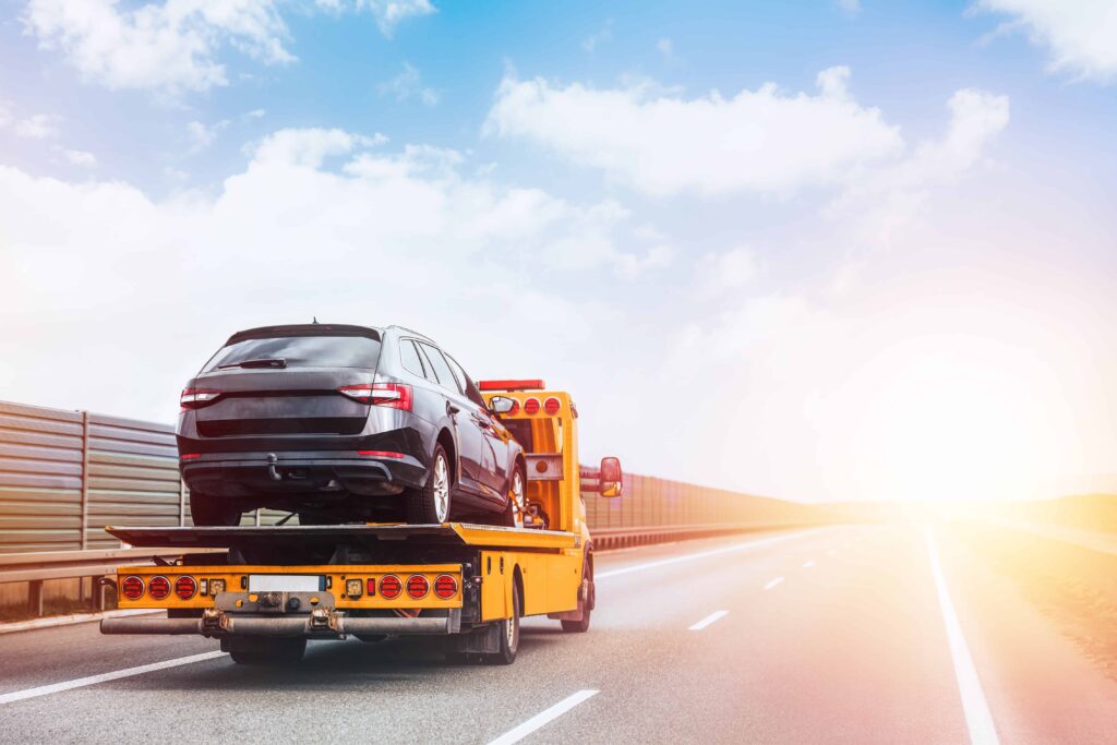 Tow truck carrying a broken-down car on highway under bright sky after vehicle accident.